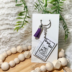 May include: A purple tassel keychain with a white card featuring a tarot card design. The card is the "XIX The Reader" and has a black and white illustration of two skulls with a book and a candle. The text "Dark Romance" is printed on the card.