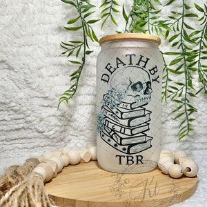 May include: White frosted glass jar with a bamboo lid. The jar has a black and white graphic of a skull with a floral design and a stack of books. The text "Death by TBR" is printed on the jar.