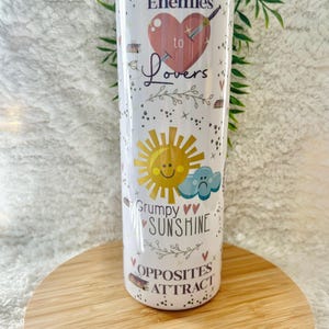 May include: White stainless steel tumbler with a colorful design featuring a heart with a sword through it, a sun and a cloud, and the text "Enemies to Lovers", "Grumpy Sunshine", and "Opposites Attract".
