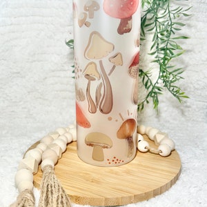 May include: A frosted glass tumbler with a lid, featuring a whimsical design of various mushrooms in shades of brown, pink, and white. The tumbler is sitting on a wooden coaster.
