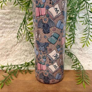 May include: A tall, cylindrical tumbler with a repeating pattern of open books, potted plants, roses, and gargoyles in shades of pink, blue, and gray on a taupe background. The tumbler is made of stainless steel.