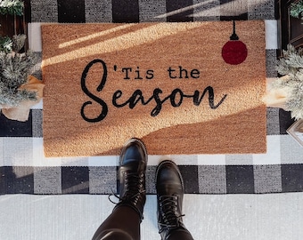 May include: A brown doormat with the text "'Tis the Season" in black cursive lettering and a red ornament hanging from the top. The doormat is on a black and white checkered surface. A person is wearing black pants and black boots.