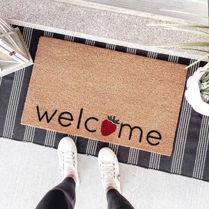 May include: A brown coir doormat with the word "welcome" in black text with a red strawberry graphic in the center of the word.