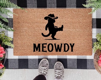 May include: A brown coir doormat with a black silhouette of a cat wearing a cowboy hat and boots. The text "MEOWDY" is printed in black below the cat.