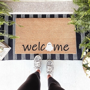 May include: A brown coir doormat with black and white striped border. The doormat has the text "welcome" in black with a white ghost in the middle of the word.