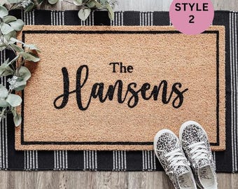 May include: A rectangular doormat with the text "The Hansens" in black script. The mat is bordered with a black frame and sits on a black and white striped rug. A pair of leopard print sneakers are placed in front of the mat.