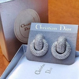 Può includere: Un paio di orecchini d'argento con un design circolare e una montatura in pietre trasparenti. Gli orecchini sono in una scatola con le parole "Christian Dior" stampate sulla scatola.