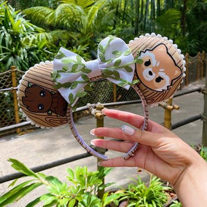 May include: A headband featuring two circular ears with a light brown wood grain pattern. The left ear displays a brown tree-like character, while the right ear shows a tan raccoon-like character. A white bow with green vine accents sits in the center.