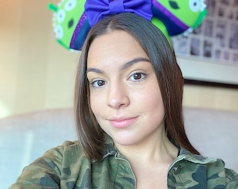 Puede incluir: Orejas de ratón con temática alienígena verde con tres ojos y un lazo morado. Las orejas están en una diadema. La persona de la imagen lleva una chaqueta de camuflaje y una camisa negra.