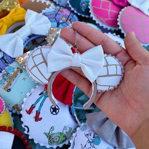 May include: White fabric Minnie Mouse ears with a brown and white concha design. The ears have a white bow and a silver metal key chain attached.