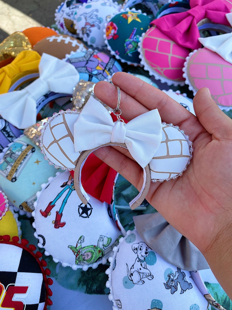 May include: White fabric Minnie Mouse ears with a brown and white concha design. The ears have a white bow and a silver metal key chain attached.