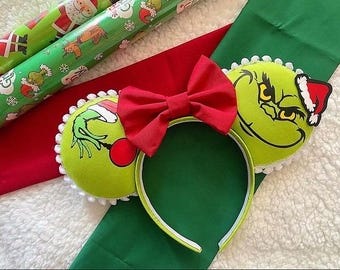 May include: Green Grinch-themed Minnie Mouse ears with a red bow and white pom-poms. The Grinch is wearing a Santa hat.