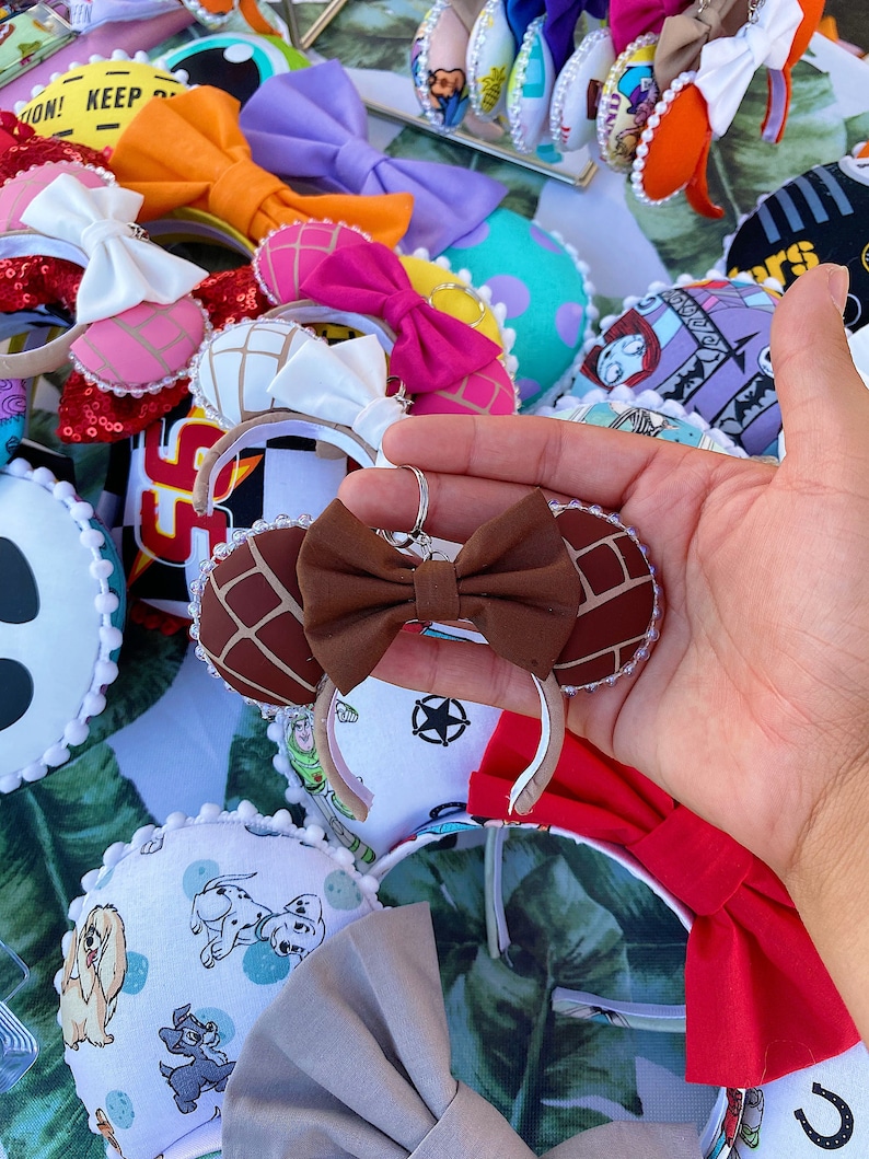 May include: A collection of handmade mouse ears with various designs, including a brown and white Mickey Mouse ear headband with a brown bow, a pink and white Mickey Mouse ear headband with a white bow, and a white and blue Mickey Mouse ear headband with a blue bow.