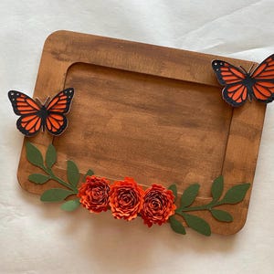 May include: A rectangular wooden frame featuring two orange and black monarch butterfly cutouts attached to the top corners and three orange paper flowers with green paper leaves arranged along the bottom edge of the frame.