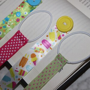 May include: Four ribbon bookmarks with elastic loops and decorative buttons. The ribbons feature patterns of flowers, polka dots, and ice cream treats. The bookmarks are placed on an open book.