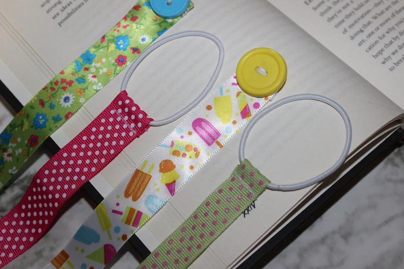 May include: Four ribbon bookmarks with elastic loops and decorative buttons. The ribbons feature patterns of flowers, polka dots, and ice cream treats. The bookmarks are placed on an open book.