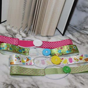 May include: Four colorful ribbon bookmarks with elastic bands and decorative buttons. The ribbons feature patterns like polka dots, floral designs, and ice cream graphics. A book is partially visible in the background.