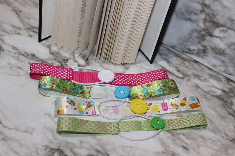 May include: Four colorful ribbon bookmarks with elastic bands and decorative buttons. The ribbons feature patterns like polka dots, floral designs, and ice cream graphics. A book is partially visible in the background.