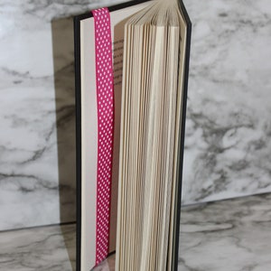 May include: A hardcover book with a black cover and red top edge, partially open to reveal the pages. A pink ribbon with white polka dots serves as a bookmark, adding a decorative touch to the reading material.