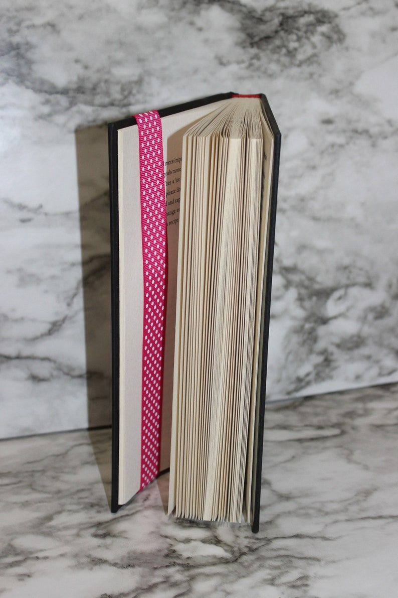 May include: A hardcover book with a black cover and red top edge, partially open to reveal the pages. A pink ribbon with white polka dots serves as a bookmark, adding a decorative touch to the reading material.