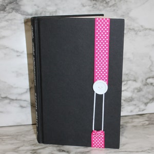 May include: A black book with a pink polka dot ribbon bookmark. The ribbon is secured with a white button and string. The book's title and author are visible on the spine.