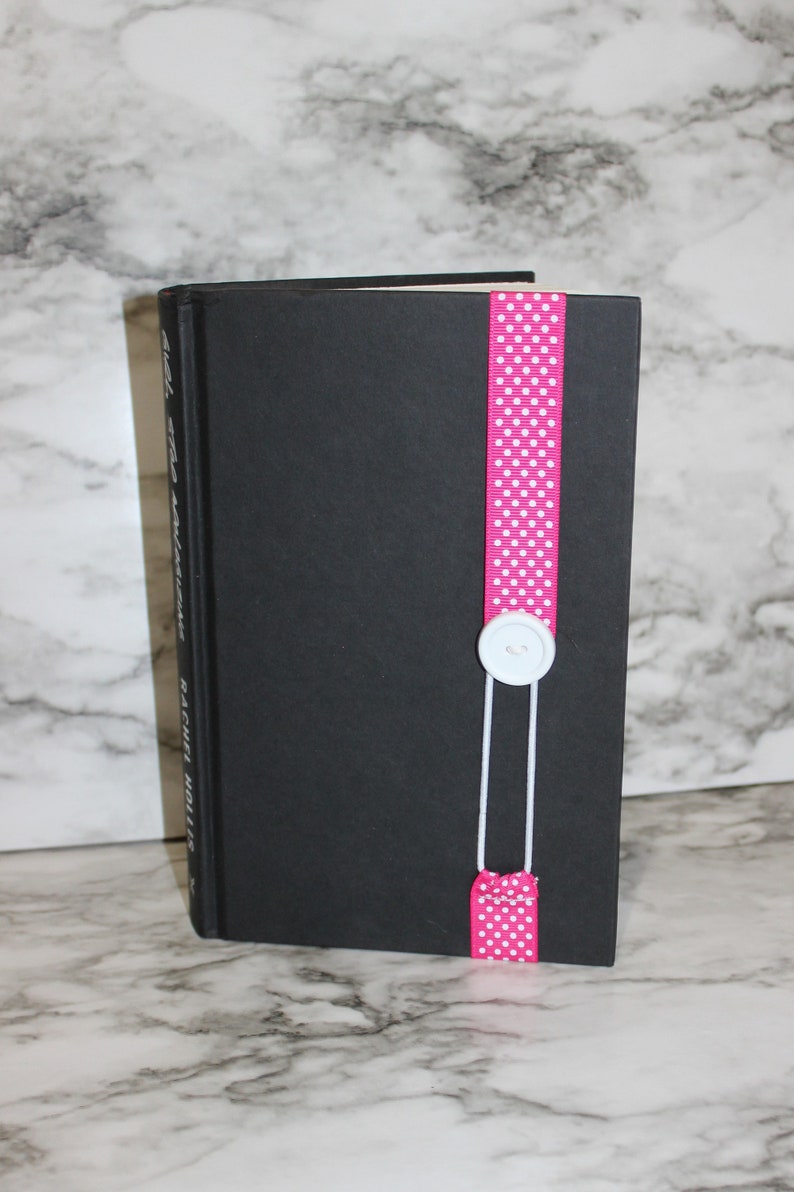 May include: A black book with a pink polka dot ribbon bookmark. The ribbon is secured with a white button and string. The book's title and author are visible on the spine.