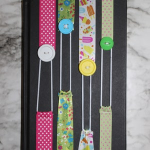 May include: A black book with colorful ribbon bookmarks. The ribbons are pink with white polka dots, green with floral patterns, and white with ice cream designs. Each ribbon has a button attached.