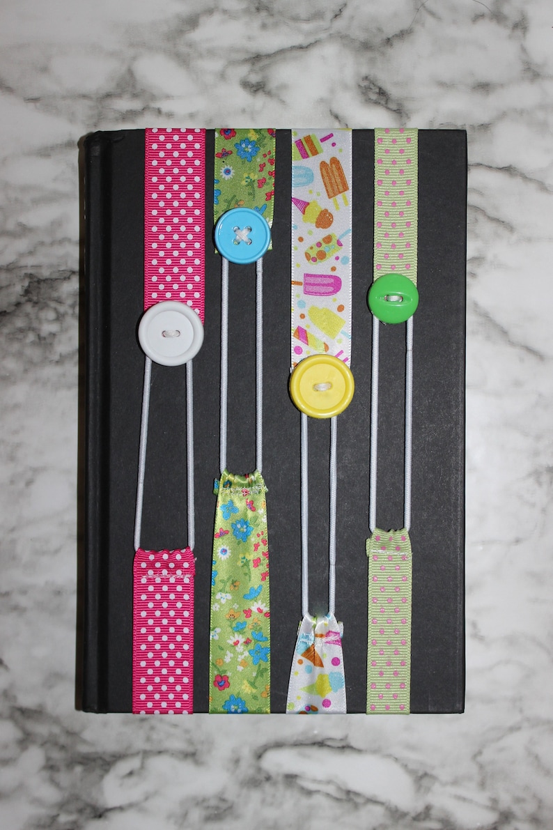 May include: A black book with colorful ribbon bookmarks. The ribbons are pink with white polka dots, green with floral patterns, and white with ice cream designs. Each ribbon has a button attached.
