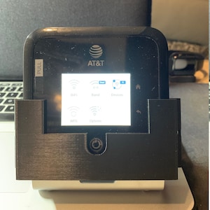 May include: A black AT&T mobile hotspot with a screen displaying WiFi, Band, Devices, WPS, and Options. The hotspot is mounted on a black stand.