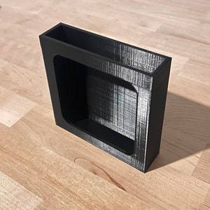 May include: A black plastic organizer with a square opening. The organizer is designed to hold small items, such as pens, pencils, or other office supplies.
