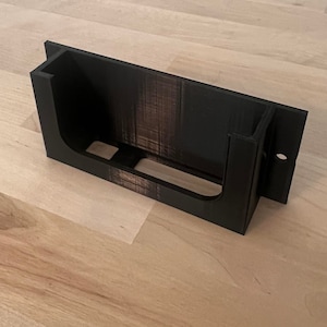 May include: A black plastic wall mount with a rectangular opening for holding a device. The mount has a small hole on the side for attaching a screw.
