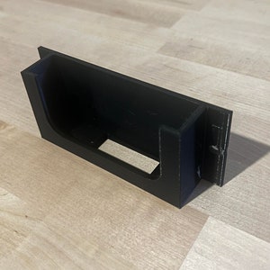 May include: A black plastic wall mount with a rectangular opening for holding a small device.