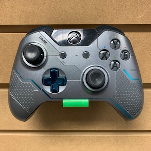 May include: A gray and blue Xbox controller with a green wall mount. The controller has a UNSC logo on the front.