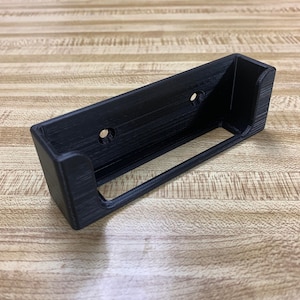 May include: A black plastic wall mount with two holes for screws. The mount has a curved lip on the front and back to hold an object in place.