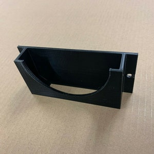 May include: A black plastic wall mount with a curved opening, designed to hold a small object. The mount has a small hole on the side for mounting.
