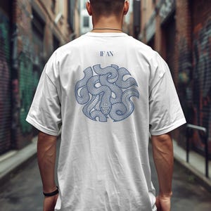 Op de afbeelding: Een wit t-shirt met korte mouwen en een blauwe afbeelding op de achterkant. De afbeelding toont een cirkelvormige opstelling van verstrengelde slangen met een rasterpatroon op hun schubben. Boven het cirkelvormige ontwerp staat de tekst IF AN in het blauw.