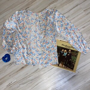 May include: A blue and white floral print long-sleeved blouse with a tie at the neck. The blouse is laid flat on a light wood surface. A blue scrunchie and a vinyl record album titled "Peter Paul and Mary In The Wind" are also visible in the image.