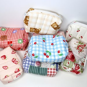 May include: A collection of quilted fabric pouches in various patterns. Designs include teddy bears, cherries, strawberries, and floral prints. The pouches are in shades of red, pink, blue, and white, with a mix of square and rectangular shapes.