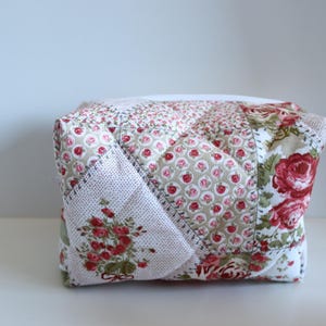 May include: A quilted fabric box featuring a patchwork design with floral patterns in red, pink, and green on a white background. The box has a square shape and is likely used for storage or decoration.