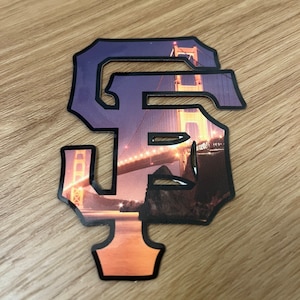 Könnte beinhalten: Ein schwarz-weißer Aufkleber mit einer Grafik des Logos der San Francisco Giants. Das Logo ist ein stilisiertes "SF" mit der Golden Gate Bridge im Hintergrund.