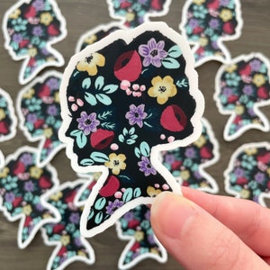 May include: A sticker featuring a black silhouette of a person's head with a floral pattern in shades of red, purple, yellow, and blue on a black background.