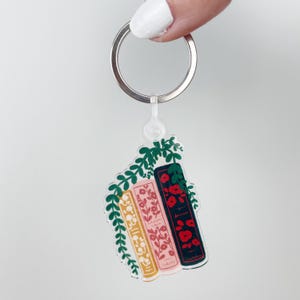 May include: A clear acrylic keychain featuring three stacked books with floral designs in yellow, pink, and dark red. Green leaves surround the books. The keychain has a silver ring.