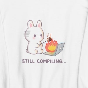 May include: White sweatshirt with a cartoon bunny toasting a marshmallow over a burning laptop. The text "STILL COMPILING..." is below the graphic. The sweatshirt has a crew neck and long sleeves.