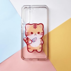 May include: Clear phone case with a cartoon tiger illustration. The tiger is orange and white with red stripes, holding a small knife. The case has the text "bakadreams" at the bottom.