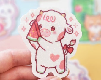 May include: A holographic sticker featuring a cartoon cow holding a watermelon slice. The cow is white with pink accents, a heart-shaped spot, and a red bow. The sticker has a sparkly, glittery effect and the text "@bakadreams".