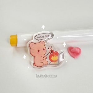 Op de afbeelding: Een heldere acryl bedel met een cartoonbeer die een marshmallow roostert boven een vuur, met de tekst "THIS IS FINE." Er is ook een kleine roze hartvormige bedel aanwezig. De bedel heeft de tekst "bakadreams" onderaan.