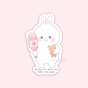 May include: A white bunny sticker with a pink blush holding a pink bottle with a cherry blossom design and a small teddy bear. The text "Being soft does not make me weak" is at the bottom.