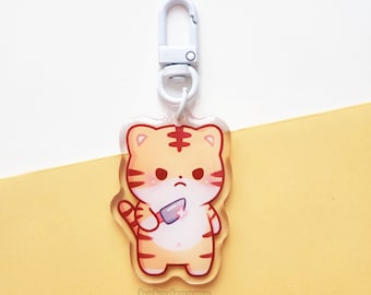 Op de afbeelding: Transparante acryl sleutelhanger met een cartoon tijger met oranje en bruine strepen, die een mes vasthoudt. De sleutelhanger heeft een witte metalen sluiting en de tekst "bakadreams" staat onderaan.