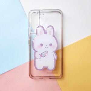 May include: Clear phone case featuring a cartoon bunny with a stern expression holding a knife. The bunny is white with pink accents and a purple outline. The text "bakadreams" is printed at the bottom of the case.