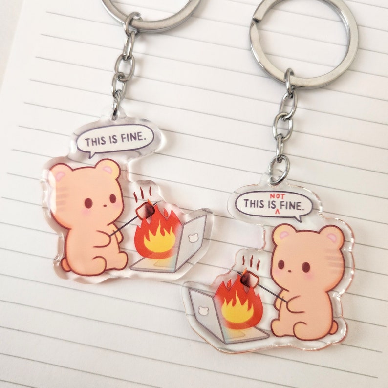 Op de afbeelding: Twee heldere acryl sleutelhangers met zilveren sleutelringen. Elke sleutelhanger toont een cartoonbeer die een marshmallow roostert boven een laptop die in brand staat. De ene zegt "THIS IS FINE" en de andere "NOT THIS IS FINE."